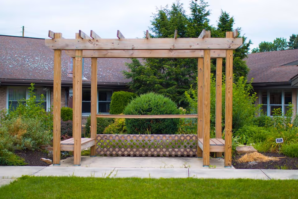 A wooden pergola structure with built-in benches on either side, situated on a concrete path surrounded by green grass and various shrubs and plants. In the background, there is a building with windows and a sloped roof partially covered by trees and bushes.