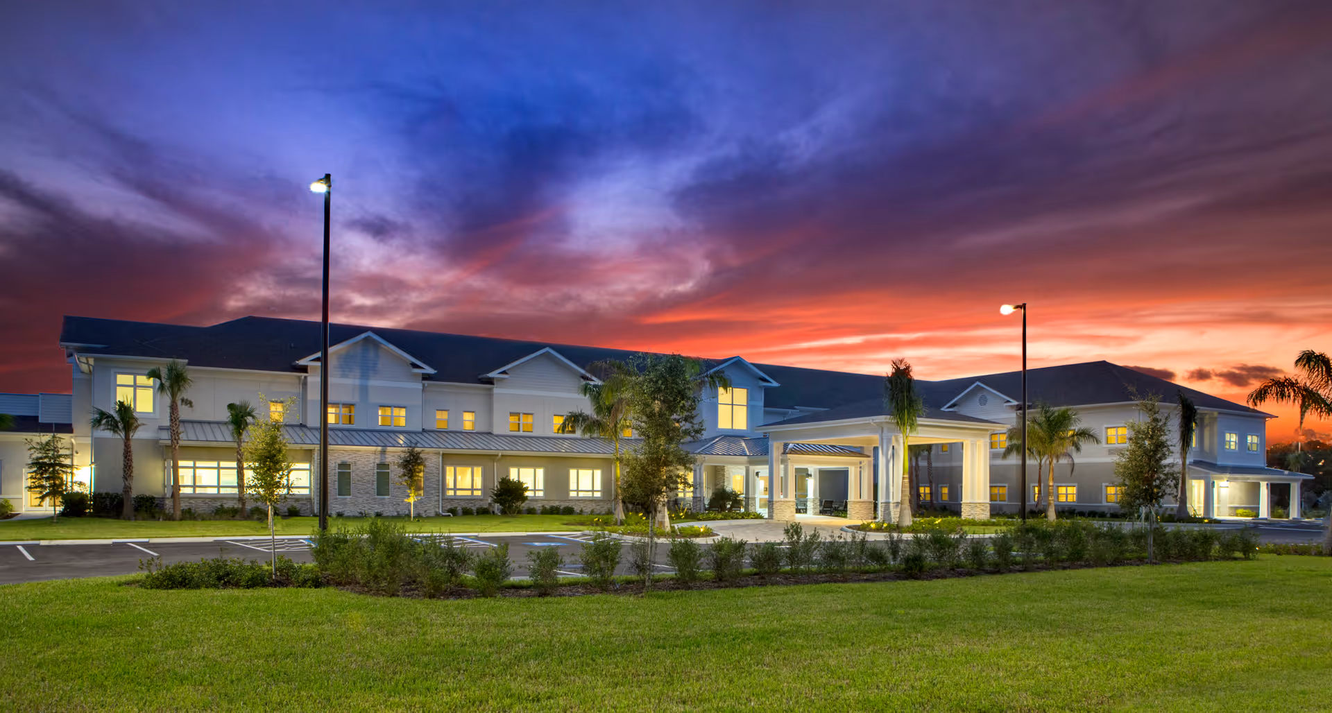Front exterior of a two-story senior living building at sunset with illuminated windows, palm trees, and a lawn.