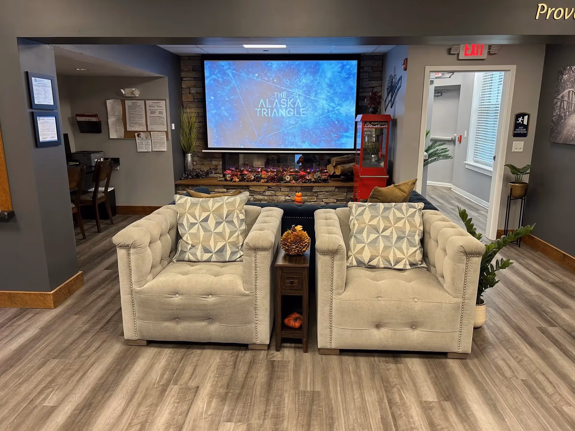 A cozy living room area with two beige armchairs featuring geometric patterned pillows, a small wooden side table with decorative pinecones, a large screen displaying 'The Alaska Triangle', a stone fireplace with logs and a red popcorn machine beside it. The room has wood flooring, gray walls, and a hallway leading to other rooms.