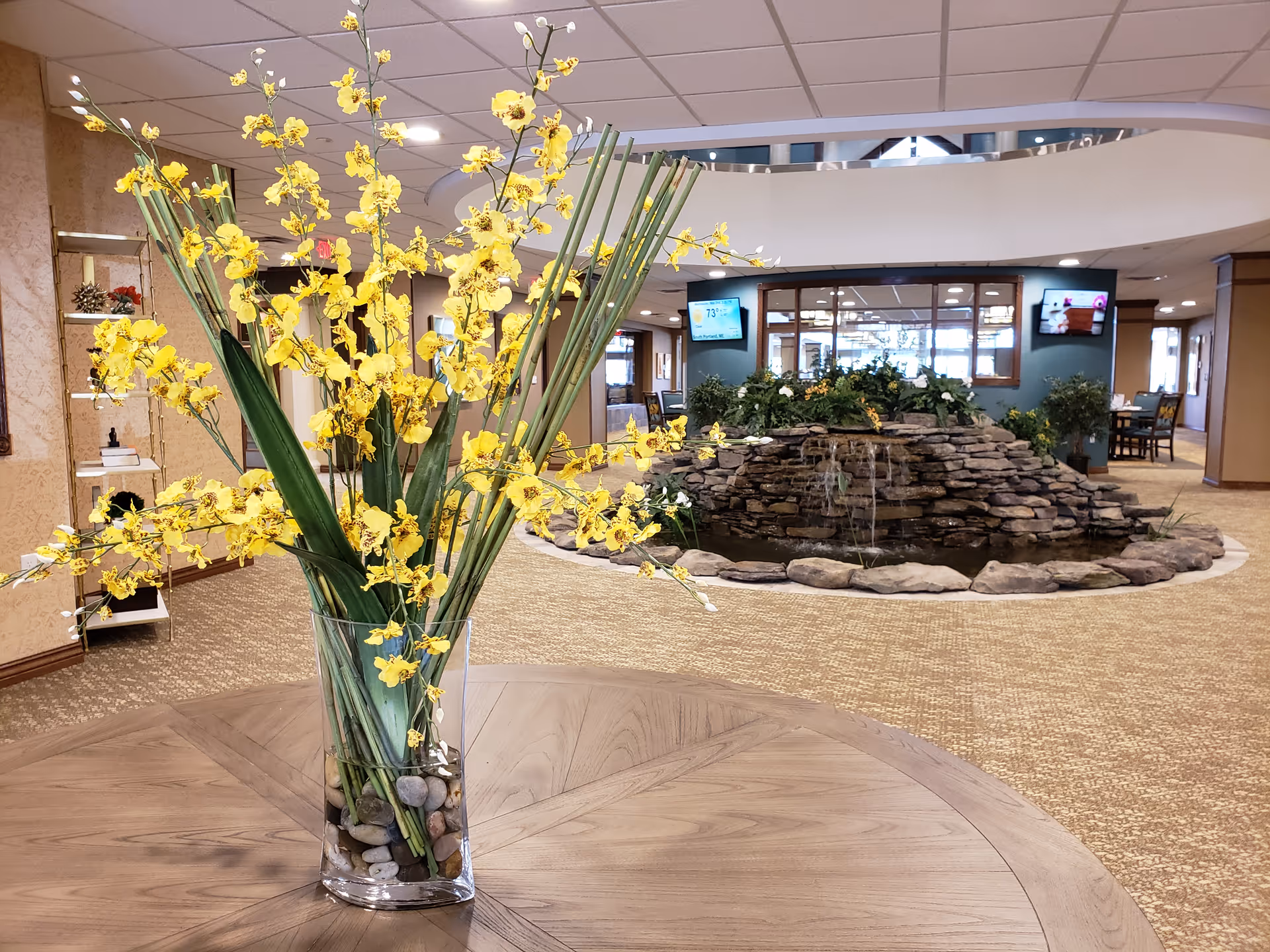 Indoor area of a retirement community featuring a round wooden table with a glass vase holding yellow flowers and green leaves. In the background, there is a decorative stone water feature surrounded by plants, with two wall-mounted televisions and seating areas visible further back.