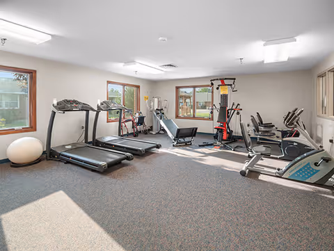 A well-lit fitness room with exercise equipment including two treadmills, stationary bikes, a multi-function weight machine, and an exercise ball. The room has carpeted flooring, beige walls, and several windows letting in natural light.