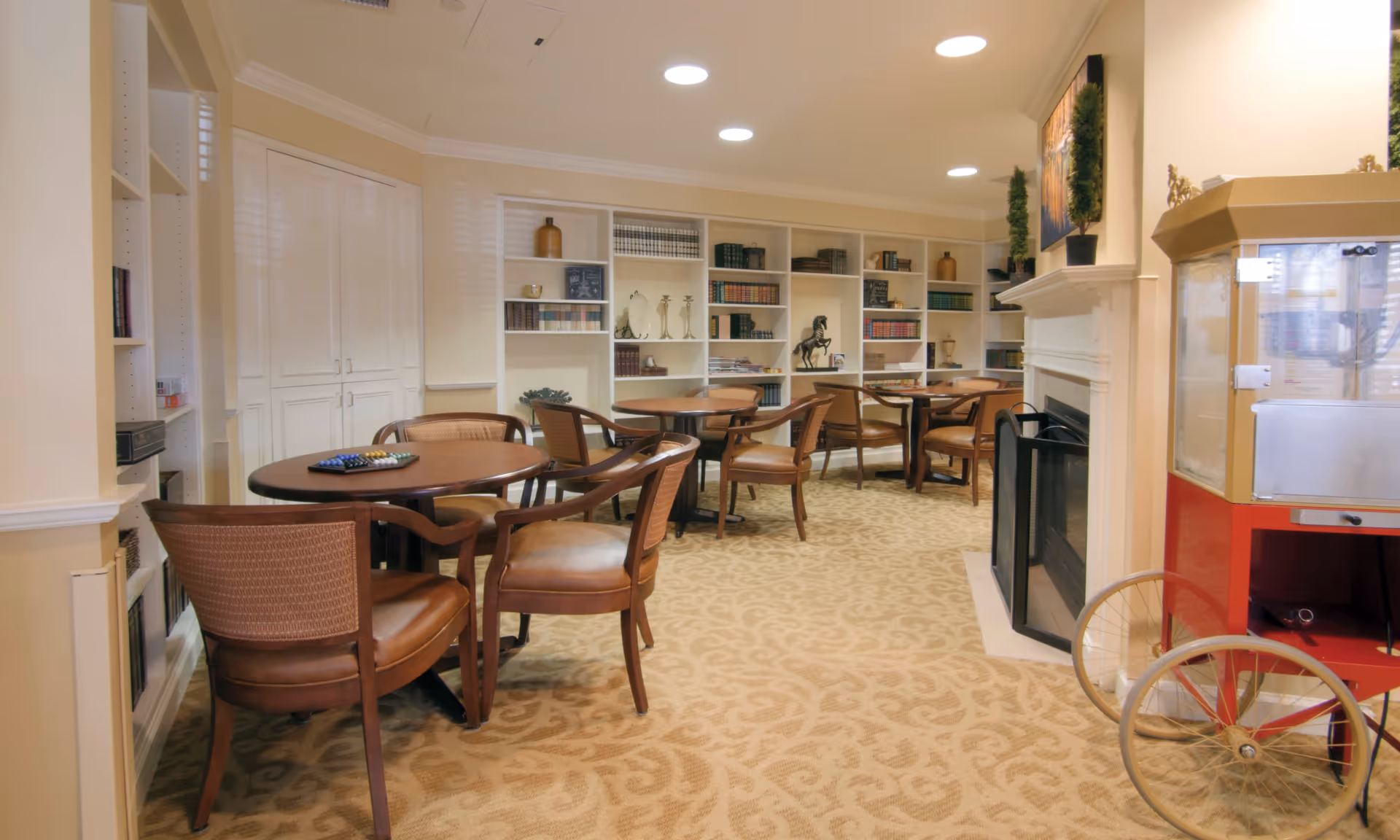 A cozy common area with round wooden tables and cushioned chairs arranged on a patterned carpet. The room features built-in white bookshelves filled with books and decorative items, a fireplace with a painting above it, and a vintage-style popcorn machine on the right side.