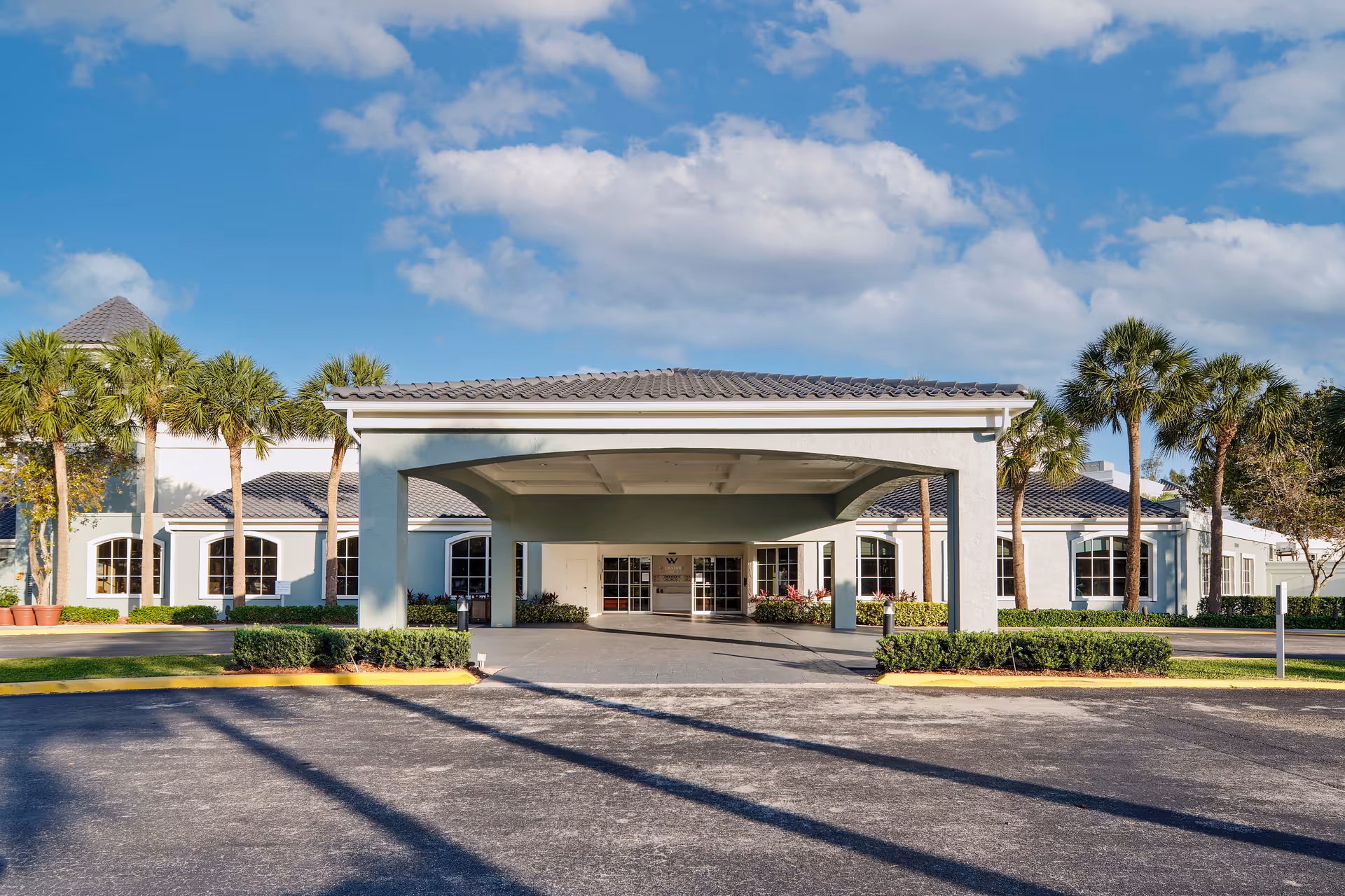 Front exterior view of a single-story building with a covered entrance, surrounded by palm trees and landscaping under a blue sky with scattered clouds.