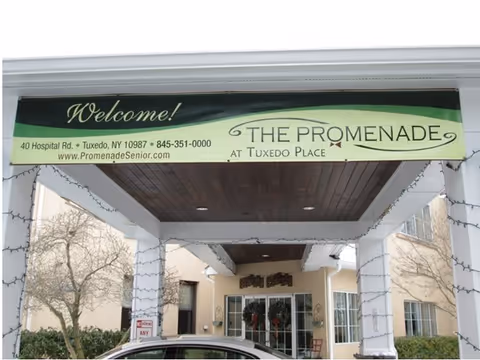 Entrance to The Promenade at Tuxedo Place senior living facility with a green and black welcome banner displaying the facility's name, address, phone number, and website. The entrance is decorated with string lights and has a covered driveway with a car parked underneath.