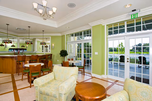 A bright and spacious common area with light green walls, featuring comfortable upholstered armchairs, wooden tables, and chairs. There is a bar area with high chairs and pendant lights, large windows and glass doors leading to an outdoor patio with seating and a view of greenery and water. The ceiling has a chandelier and recessed lighting.