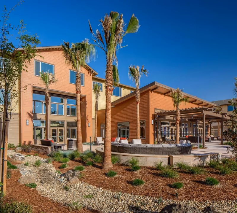 Outdoor patio area of a senior living facility with palm trees, landscaped garden beds with rocks and mulch, and modern seating arrangements under a pergola. The building has large windows and a warm-toned exterior under a clear blue sky.