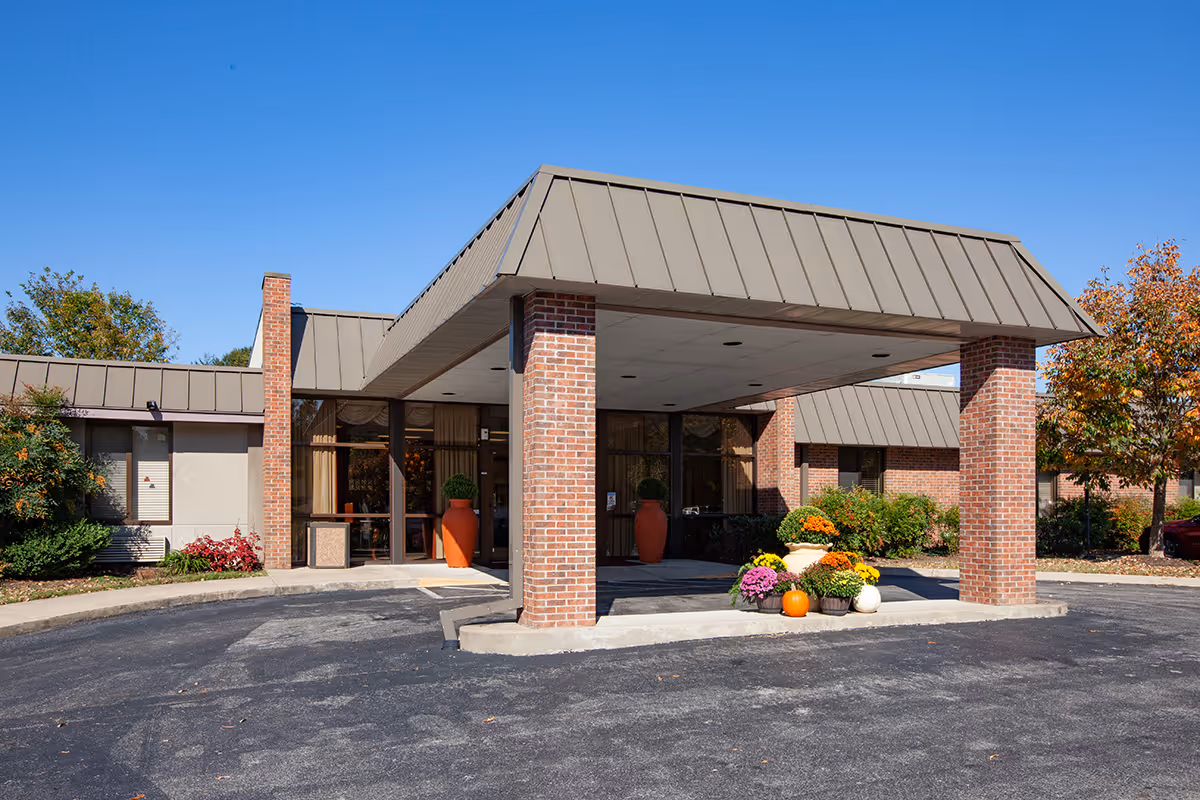 The front entrance of Alta Heights Post Acute facility featuring a covered drop-off area supported by brick columns. There are large windows and glass doors at the entrance, with decorative potted plants and flowers, including pumpkins, arranged near the entrance. The building has a brown metal roof and brick exterior walls. Trees with autumn foliage are visible on the right side under a clear blue sky.