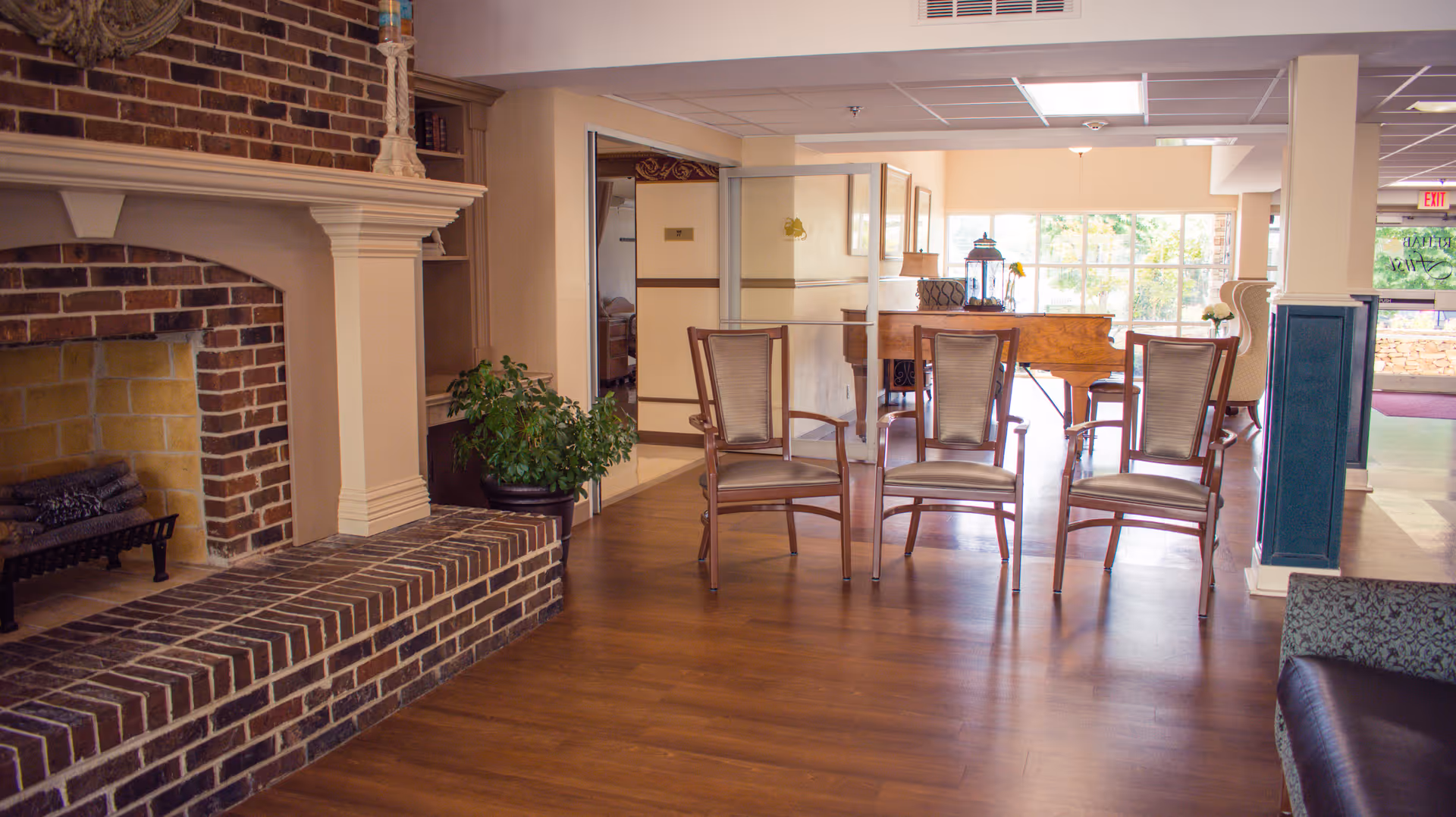 Communal lounge with a brick fireplace, a row of chairs, a piano, and large windows.