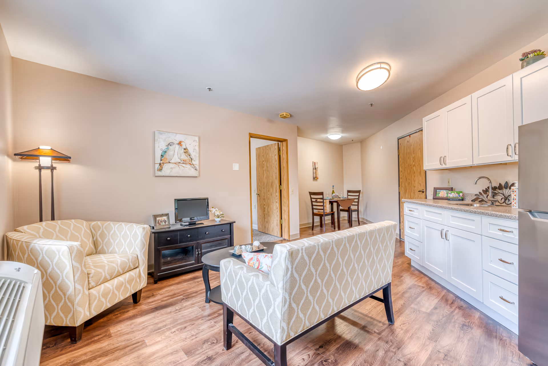 A bright and cozy senior living apartment featuring a small living area with two patterned armchairs and a loveseat around a round coffee table. A floor lamp stands in the corner next to a dark wooden TV stand with a small television and decorative items. The room has wood flooring and beige walls with a painting of two birds. To the right is a kitchenette with white cabinets, a sink, and a stainless steel refrigerator. In the background, there is a small dining area with a table and two chairs.