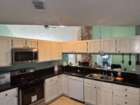 Interior view of a kitchen with white cabinets, black countertops, a stainless steel microwave, stove, and dishwasher. The kitchen has a double sink, various kitchen items on the counters, and a light green wall in the background.