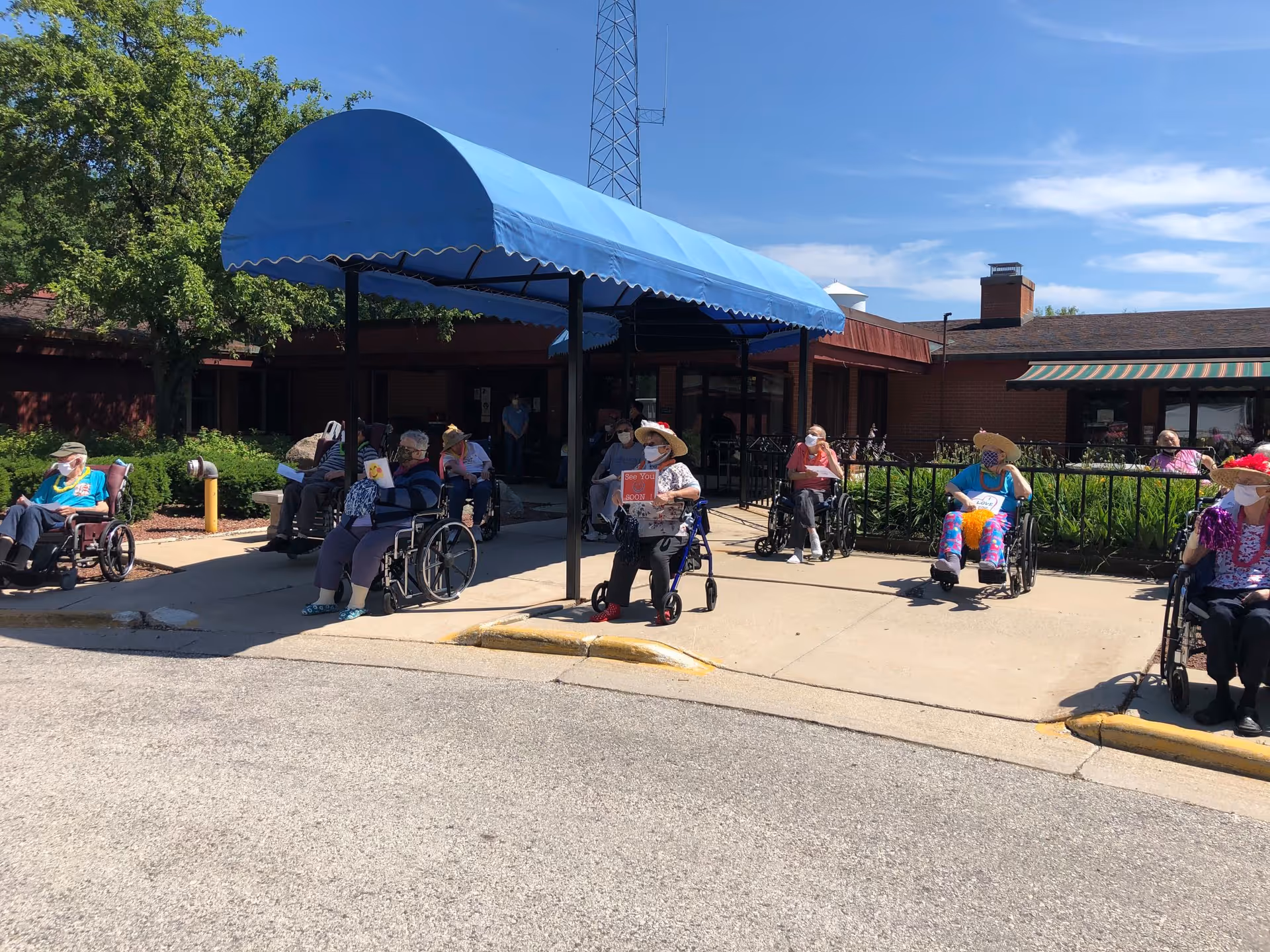A group of elderly people sitting outside a building under a blue awning. Most are in wheelchairs or using walkers, wearing face masks and colorful hats. The building has a brick exterior and there are trees and bushes around the area.