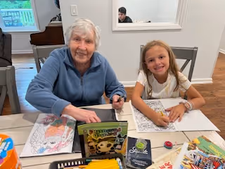 An elderly woman and a young girl sitting at a table engaged in coloring activities. They are smiling and appear to be enjoying their time together. Various coloring books, crayons, and colored pencils are spread out on the table in front of them. The background shows a piano and a window.