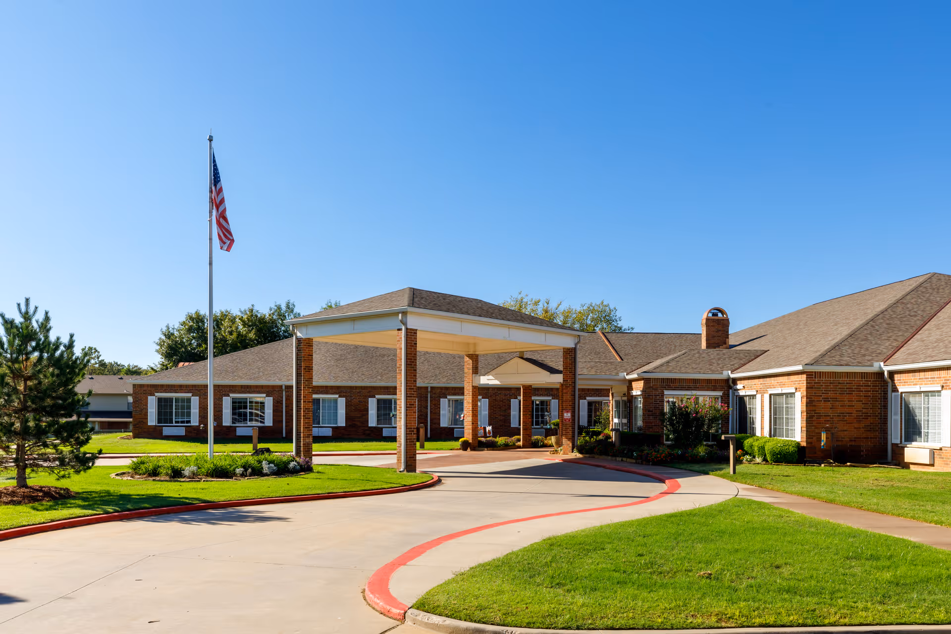 Exterior view of Rivermont Assisted Living and Memory Care building with a covered entrance, brick facade, an American flag on a flagpole, well-maintained green lawn, and clear blue sky.