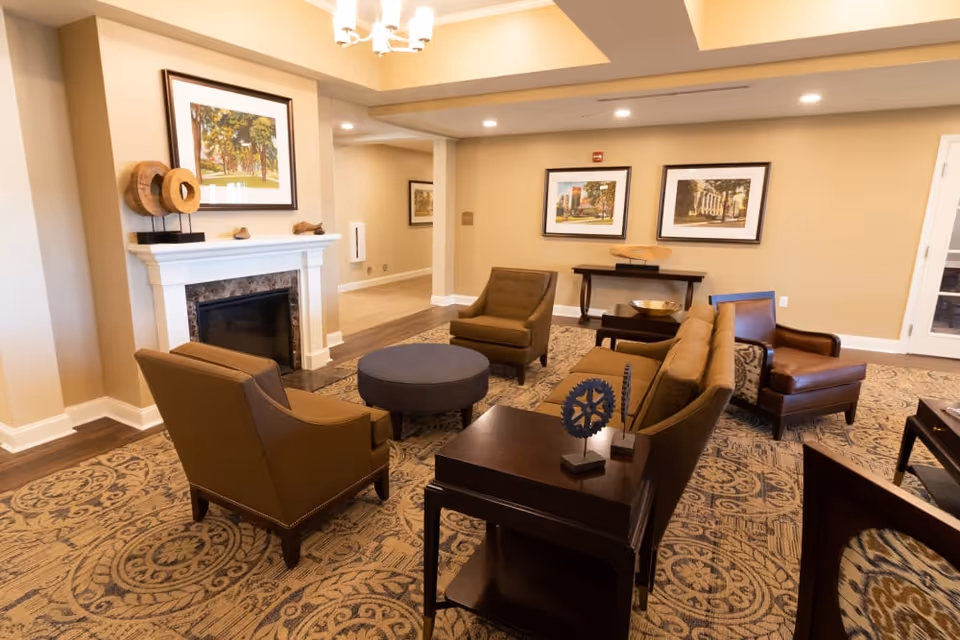 A cozy living room area with beige walls and patterned carpet. The room features a fireplace with decorative items on the mantle, a round ottoman, two brown armchairs, a brown sofa, and a leather armchair. There are framed pictures on the walls and a wooden console table with decorative bowls.