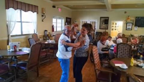 Two people dancing in a dining room of an assisted living facility while other residents sit at tables.