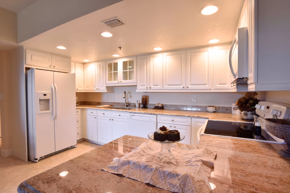 A bright kitchen with white cabinets, a side-by-side refrigerator, a built-in microwave, an electric stove, and a granite countertop island with a decorative cloth and a glass cake stand holding small cakes.