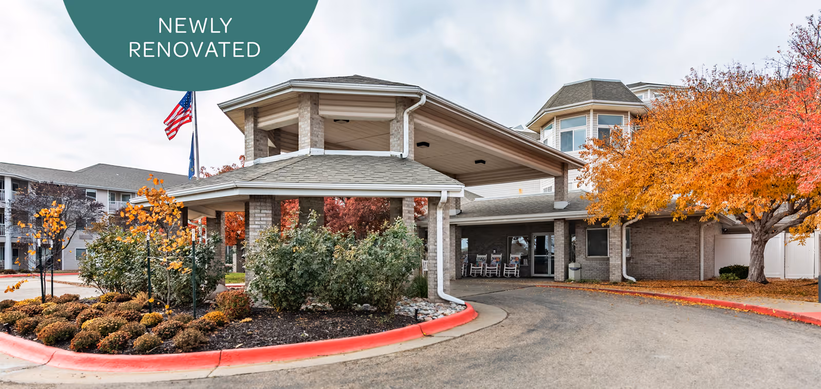 Front entrance of a senior living building with a covered porte-cochere, landscaped beds and autumn trees under a 'NEWLY RENOVATED' banner.