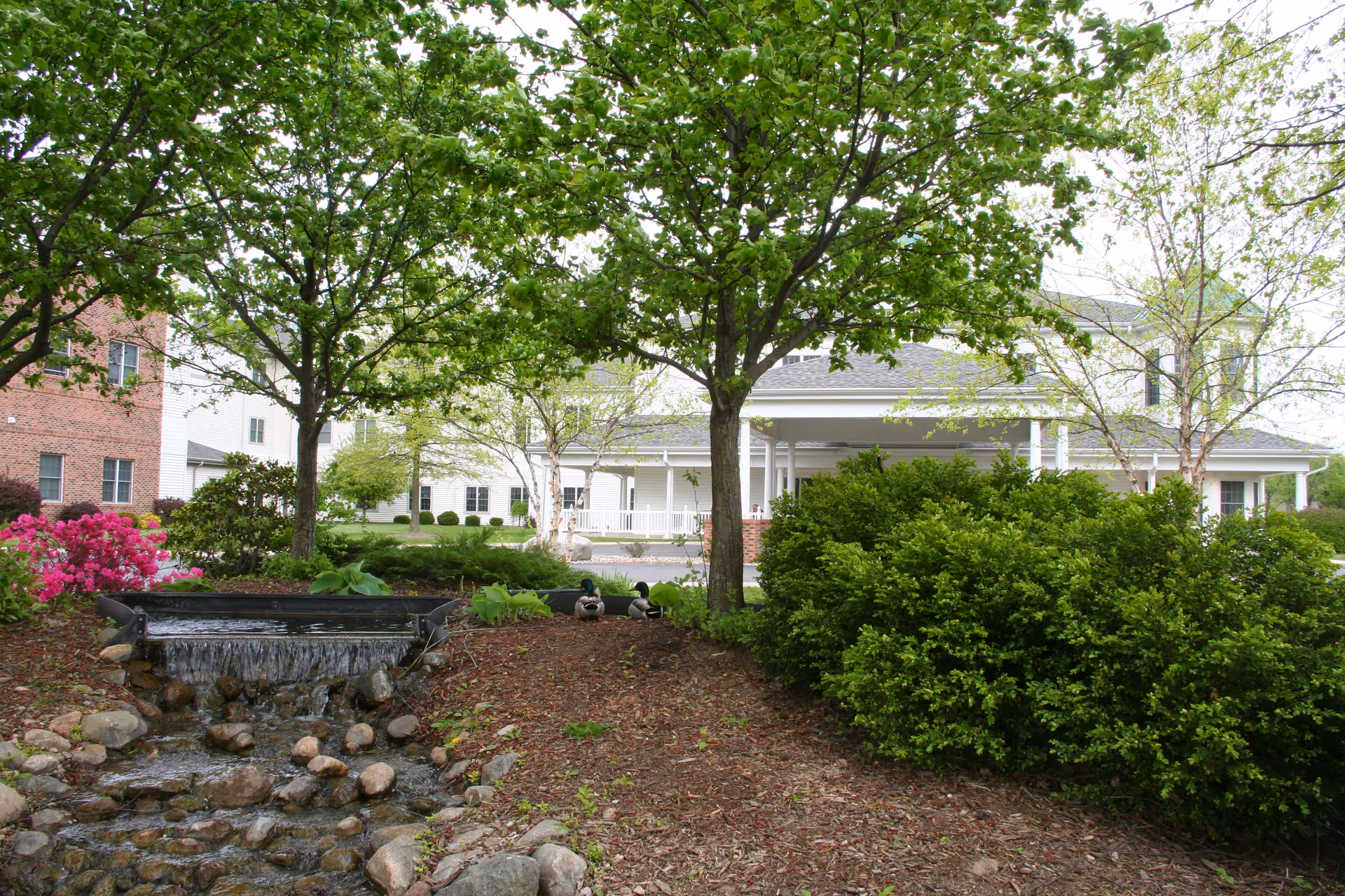 Outdoor garden area with green trees, bushes, and a small waterfall flowing over rocks. In the background, there is a white building with a covered porch and a brick building partially visible on the left.