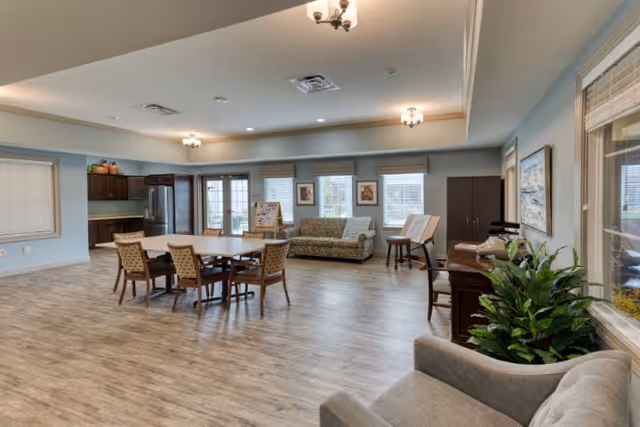 A spacious and well-lit common area in a senior living facility featuring a dining table with six chairs, a sofa near windows with framed artwork, a kitchenette with dark cabinets and stainless steel refrigerator, and additional seating including armchairs. The room has wood flooring, light blue walls, and ceiling light fixtures.