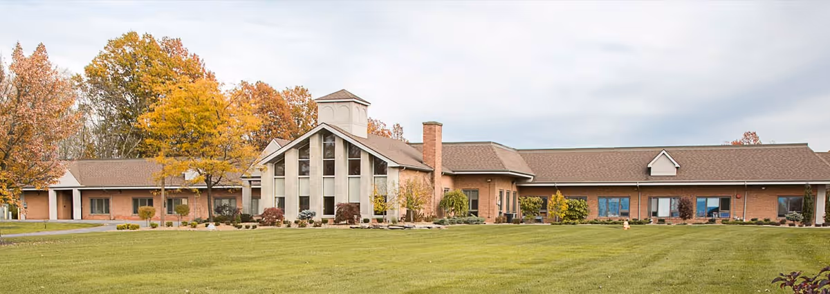 Exterior view of a single-story senior living facility building with a large green lawn in front and trees with autumn foliage in the background under a cloudy sky.