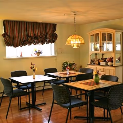 A cozy dining area with three tables, each surrounded by four black chairs. Each table has a small vase with flowers. There is a window with a striped valance, a hanging light fixture above one table, and a cream-colored cabinet with glass doors displaying decorative items in the background.