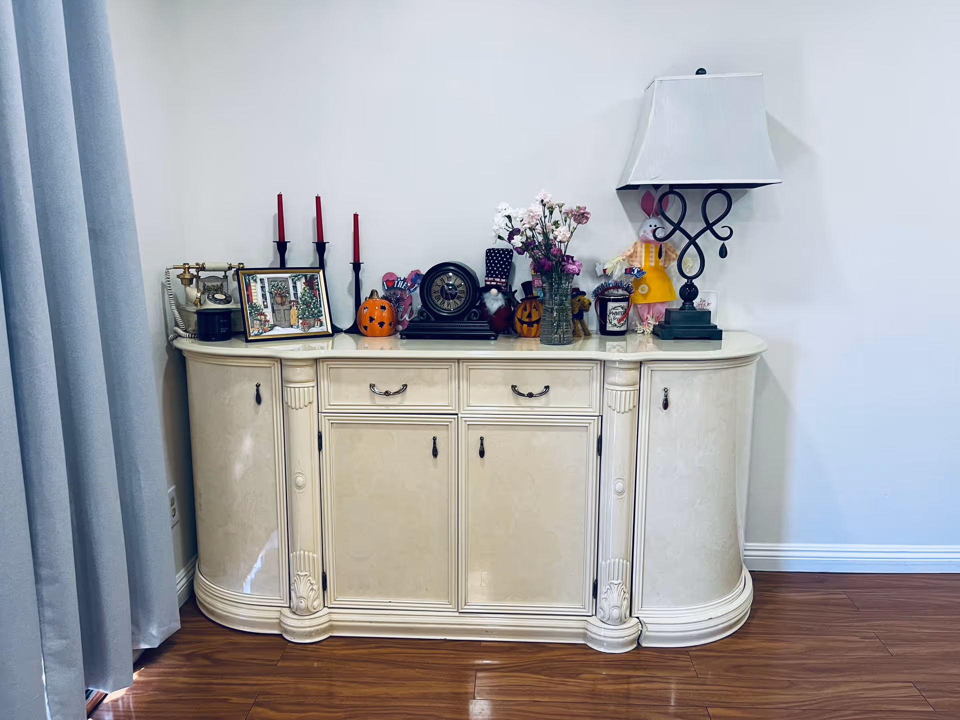 A cream-colored vintage sideboard with decorative carvings stands against a white wall on a wooden floor. On top of the sideboard are various decorative items including a vintage rotary phone, a framed picture, three red candles in black holders, two small pumpkin decorations, a clock, a vase with flowers, a candle, a bunny doll, and a table lamp with a white shade and black ornate base. To the left of the sideboard is a gray curtain.