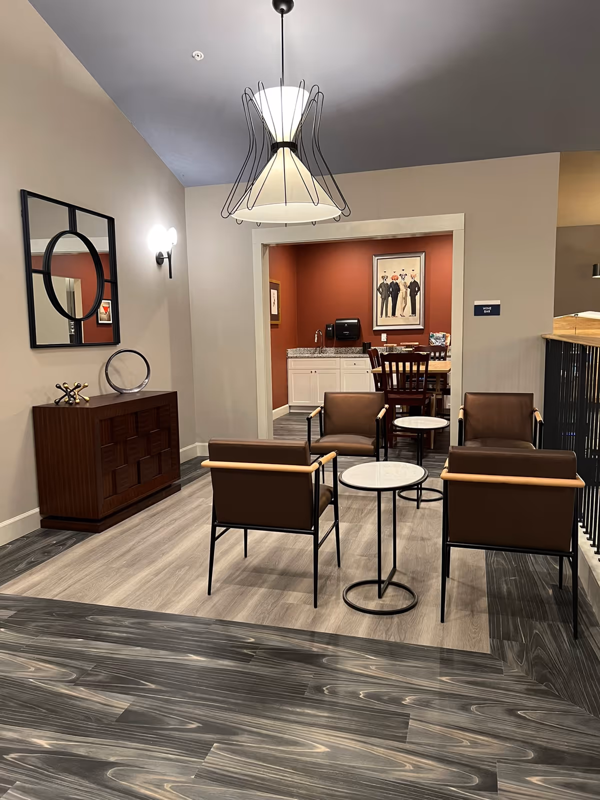 Modern lounge area with four brown armchairs around small round tables, a sideboard with mirror, and a view into a dining/kitchen alcove.
