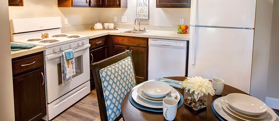 A kitchen with dark wood cabinets, a white electric stove with oven, a white dishwasher, and a white refrigerator. A round dining table is set with plates, bowls, mugs, and a small flower arrangement. The countertop has a sink and a decorative sign that reads 'Eat Drink & Give Thanks'.