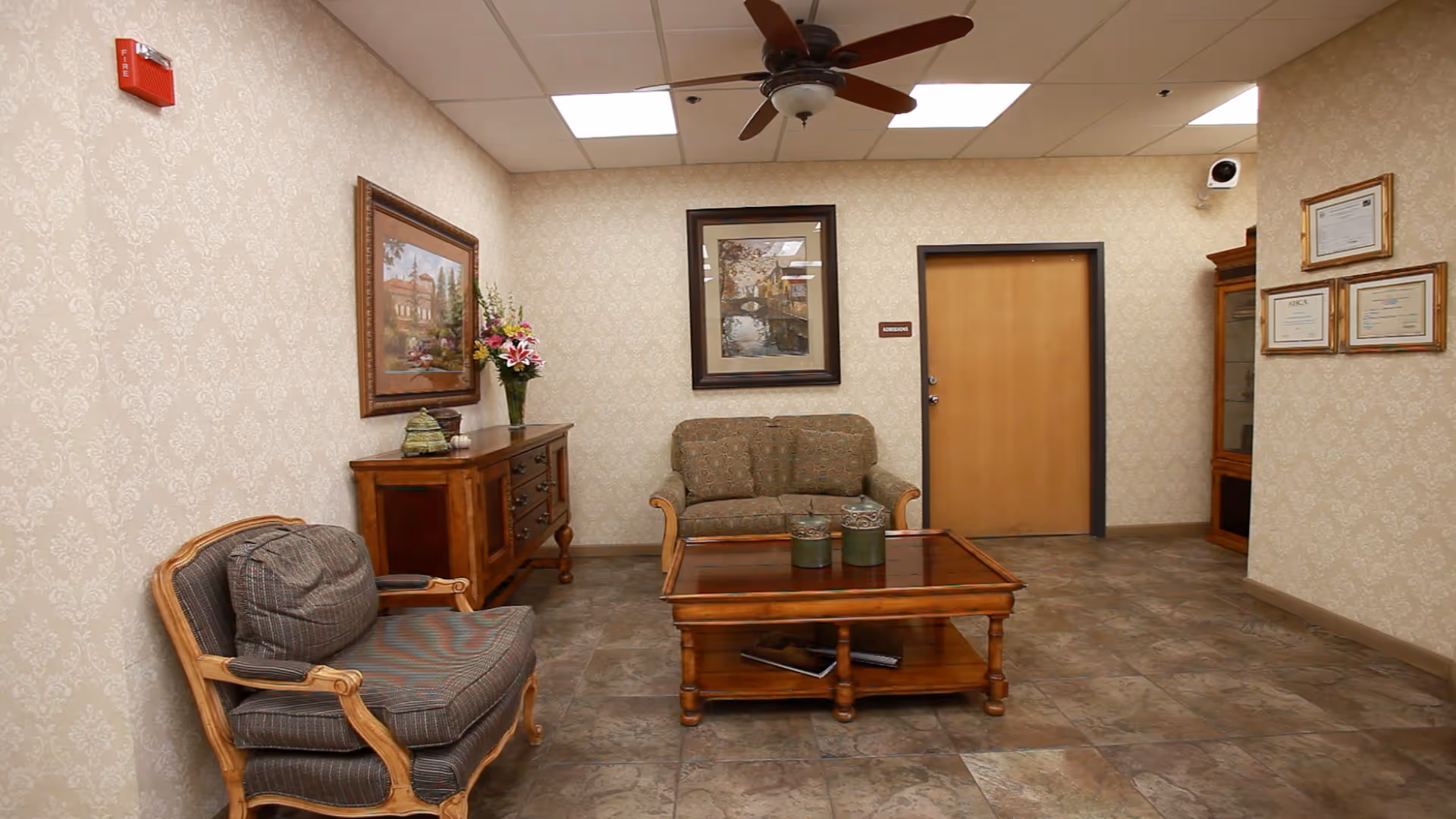 A small sitting area in a nursing center with two upholstered chairs, a wooden coffee table with decorative items, a wooden sideboard with a flower vase, framed paintings on the walls, and certificates displayed on the right wall. The room has tiled flooring, patterned wallpaper, a ceiling fan, and a closed wooden door.