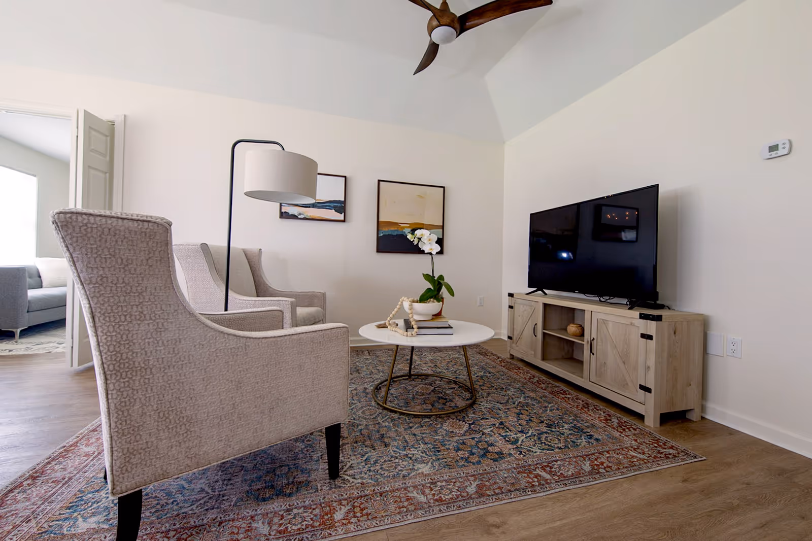 Bright living room with two beige armchairs, a round coffee table, a TV on a wooden console, and a patterned area rug.