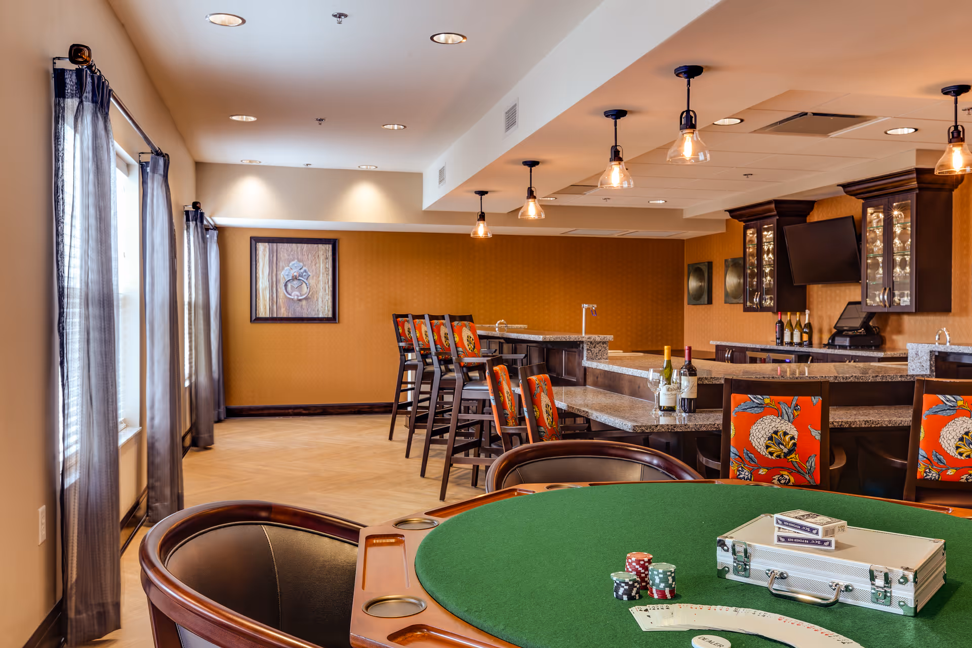 Interior view of a senior living facility's recreational and bar area featuring a green poker table with poker chips, playing cards, and a silver case. In the background, there is a bar counter with high chairs, bottles of wine, and a mounted TV. The room has warm lighting, large windows with sheer curtains, and decorative wall art.