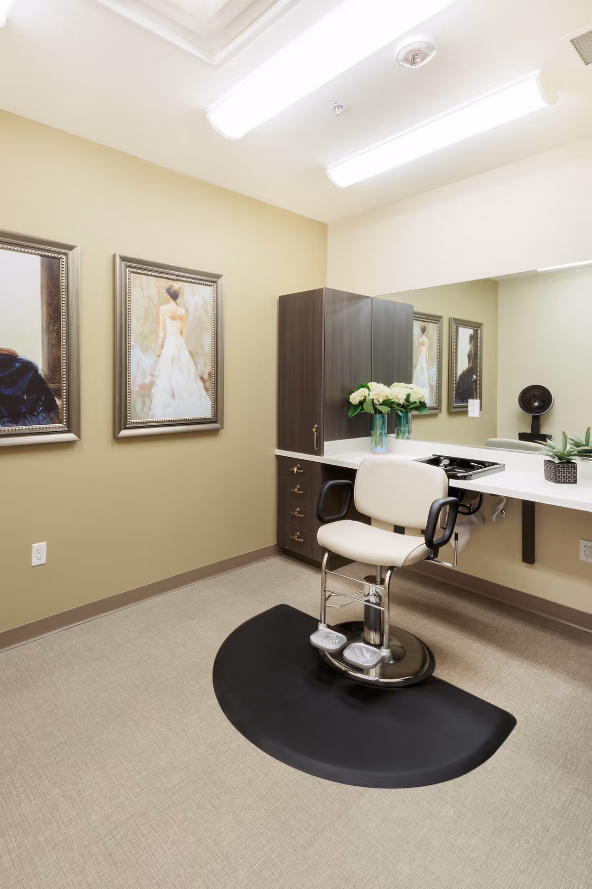 A small salon or grooming area with a white salon chair on a black mat in front of a counter with a large mirror. The counter has a vase with white flowers and a small potted plant. There are two framed paintings on the beige wall, one depicting a woman in a white dress. The room has beige walls and carpeted floor.