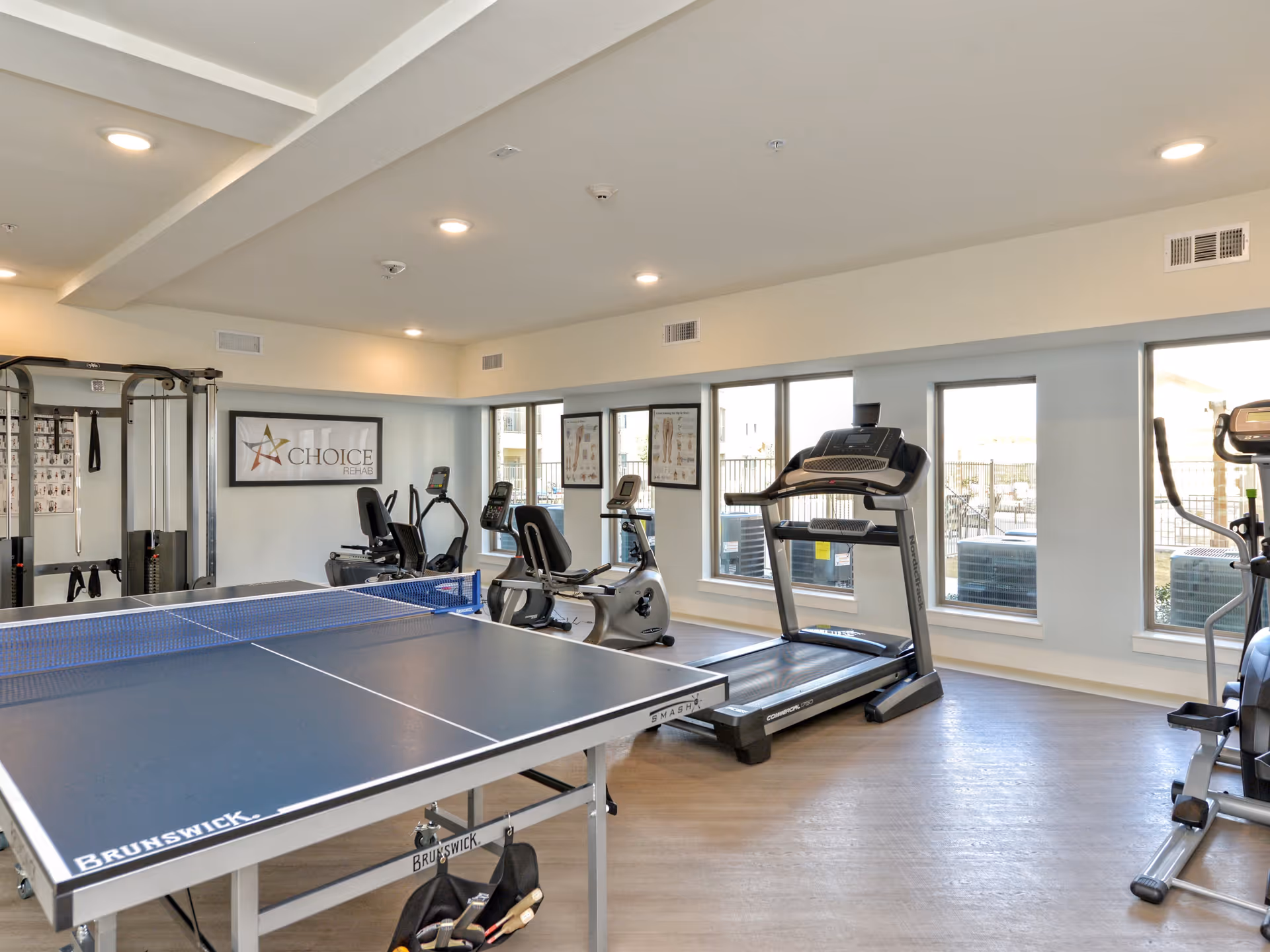 A bright fitness room with a ping-pong table in the foreground and treadmills, exercise bikes and strength equipment along the windows.