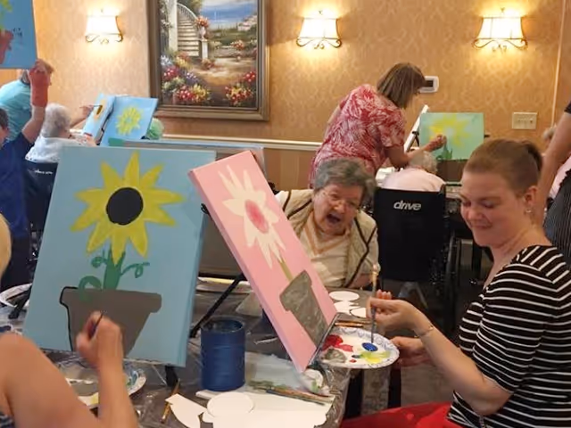 A group of elderly individuals and caregivers participating in a painting activity in a well-lit room with beige wallpaper and framed artwork. Several canvases with flower paintings are visible, and people are seated around tables, some in wheelchairs, engaging in the creative activity.