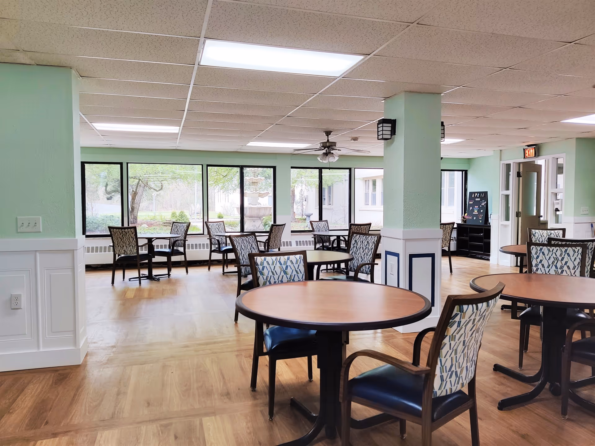 A bright dining room with multiple round wooden tables and patterned chairs. Large windows along one wall provide a view of greenery and a fountain outside. The room has light green walls, a white ceiling with fluorescent lights, and a ceiling fan. There is an exit door on the right side of the room.