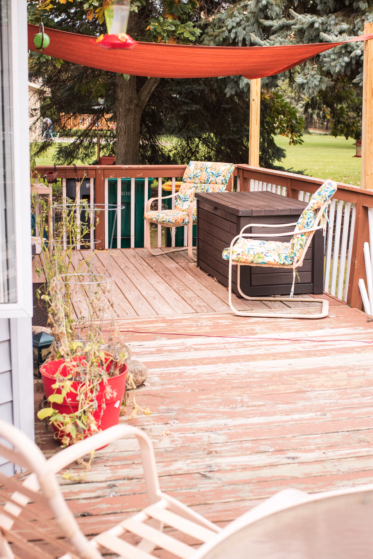 Outdoor wooden deck area with two floral cushioned chairs and a dark brown storage box. A red shade sail is stretched overhead, and there are potted plants on the deck. Trees and green grass are visible in the background.
