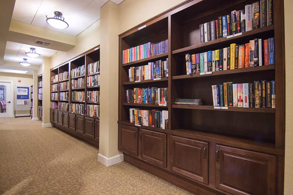 A hallway lined with built-in wooden bookshelves filled with books on the right side, beige walls, and carpeted floor. Ceiling lights illuminate the space, and a door is visible at the end of the hallway.