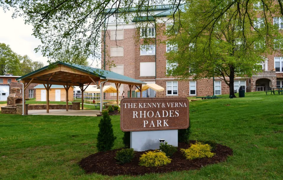 Outdoor park area with a sign reading 'The Kenny & Verna Rhoades Park' in front of a green lawn, trees, a covered pavilion with a green roof, picnic tables, and a large brick building in the background.