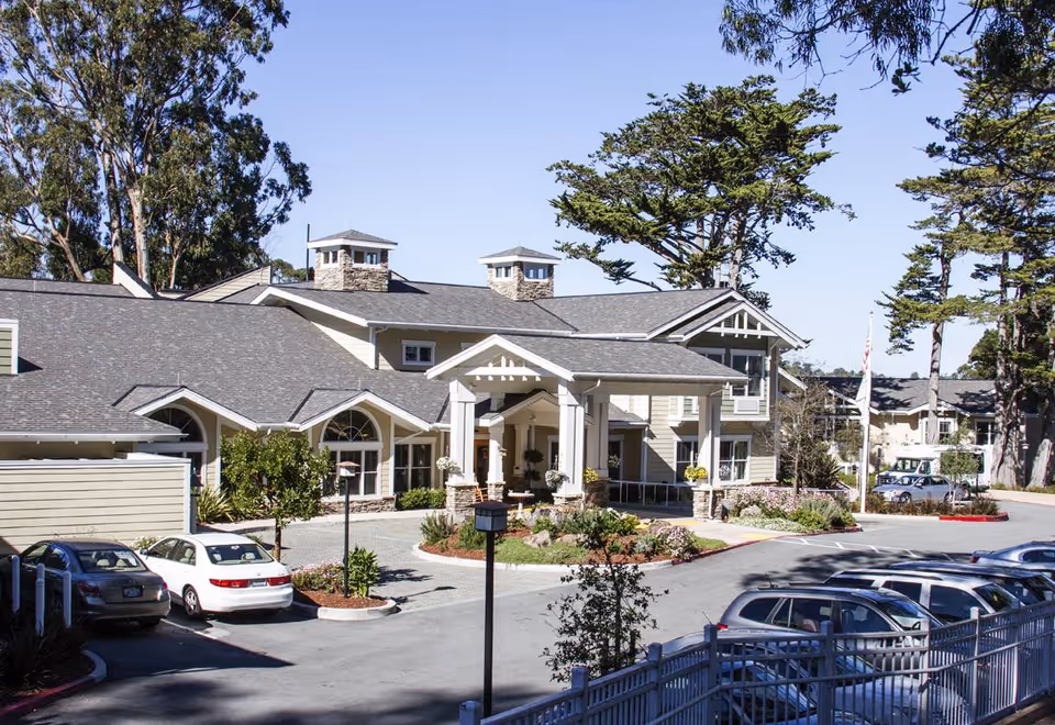 Exterior view of a senior living facility named Ivy Park at Monterey, showing a large building with a covered entrance, multiple windows, and a parking lot with several cars. Tall trees surround the property under a clear blue sky.