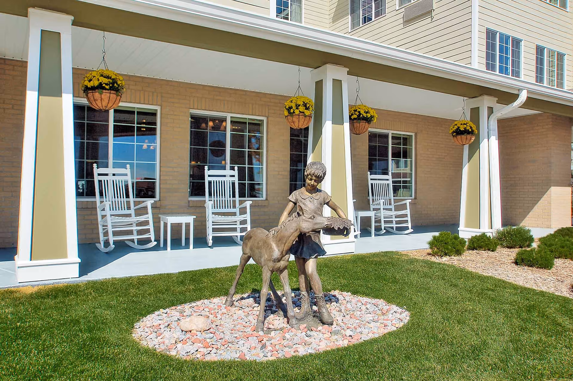Outdoor garden area in front of a building with a covered porch featuring white rocking chairs and hanging flower baskets. In the foreground, there is a bronze statue of a young girl standing next to a foal on a bed of decorative rocks surrounded by green grass.