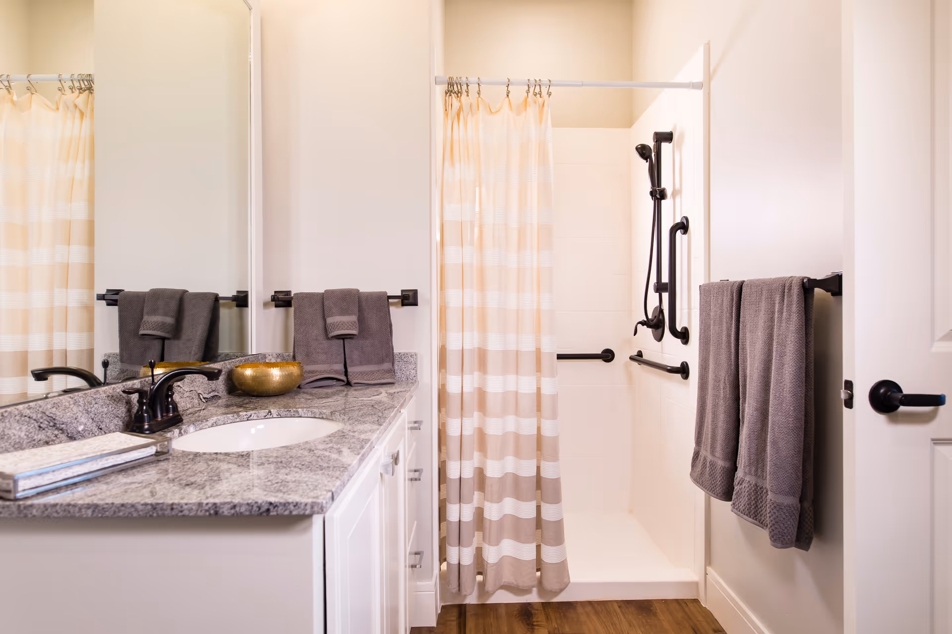 A bathroom with a granite countertop vanity featuring a sink, a faucet, and a decorative bowl. There are two gray towels hanging on a towel rack above the countertop and two more gray towels hanging on a rack on the wall near the door. The shower area has a striped beige and white curtain and multiple black grab bars installed on the walls for accessibility. The floor is wood-style, and the walls are painted white.