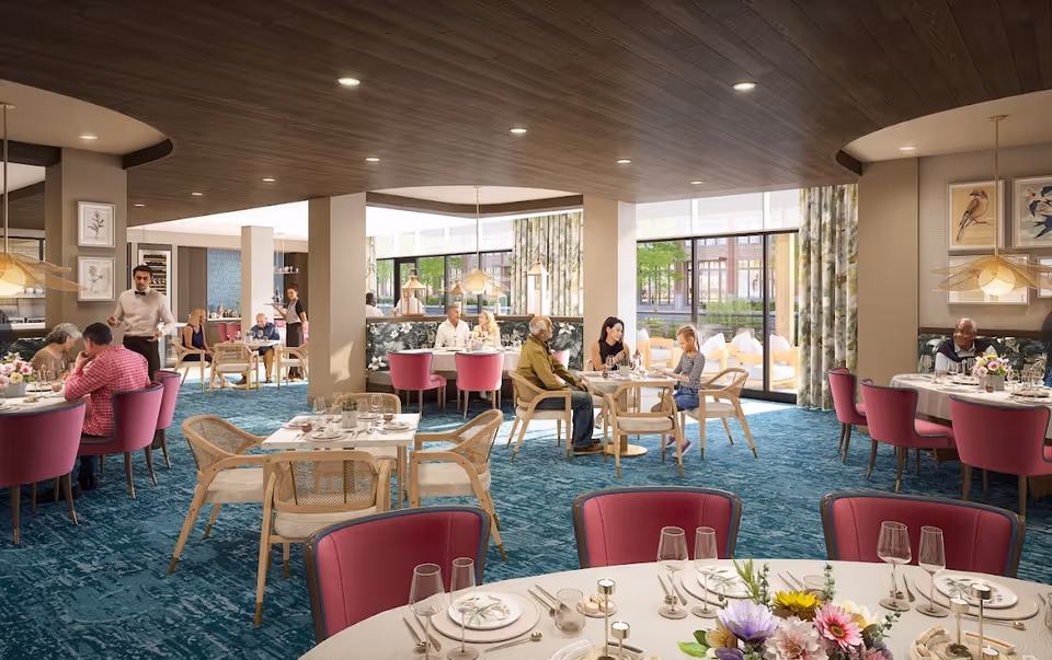 A bright and spacious dining room in a senior living facility with several tables set for meals. People of various ages are seated and engaged in conversation. The room features large windows with floral curtains, blue carpet, wooden ceiling, and modern light fixtures. Waitstaff are attending to guests.
