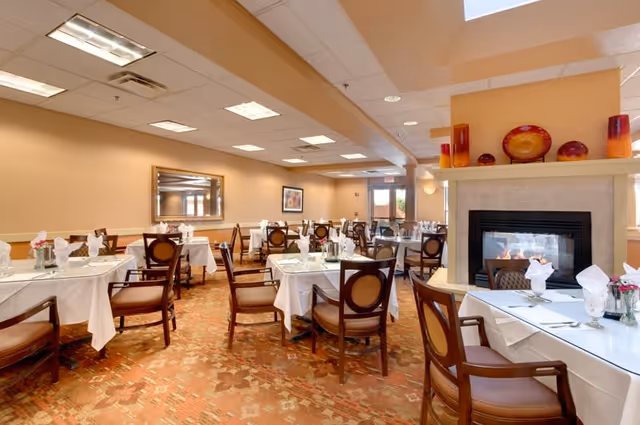 A dining room with multiple tables covered in white tablecloths, each set with glassware, napkins, and silverware. The room features wooden chairs with cushioned seats, a fireplace with decorative vases and plates on the mantel, warm beige walls, a large mirror, and framed artwork. The carpet has a patterned design in earthy tones.