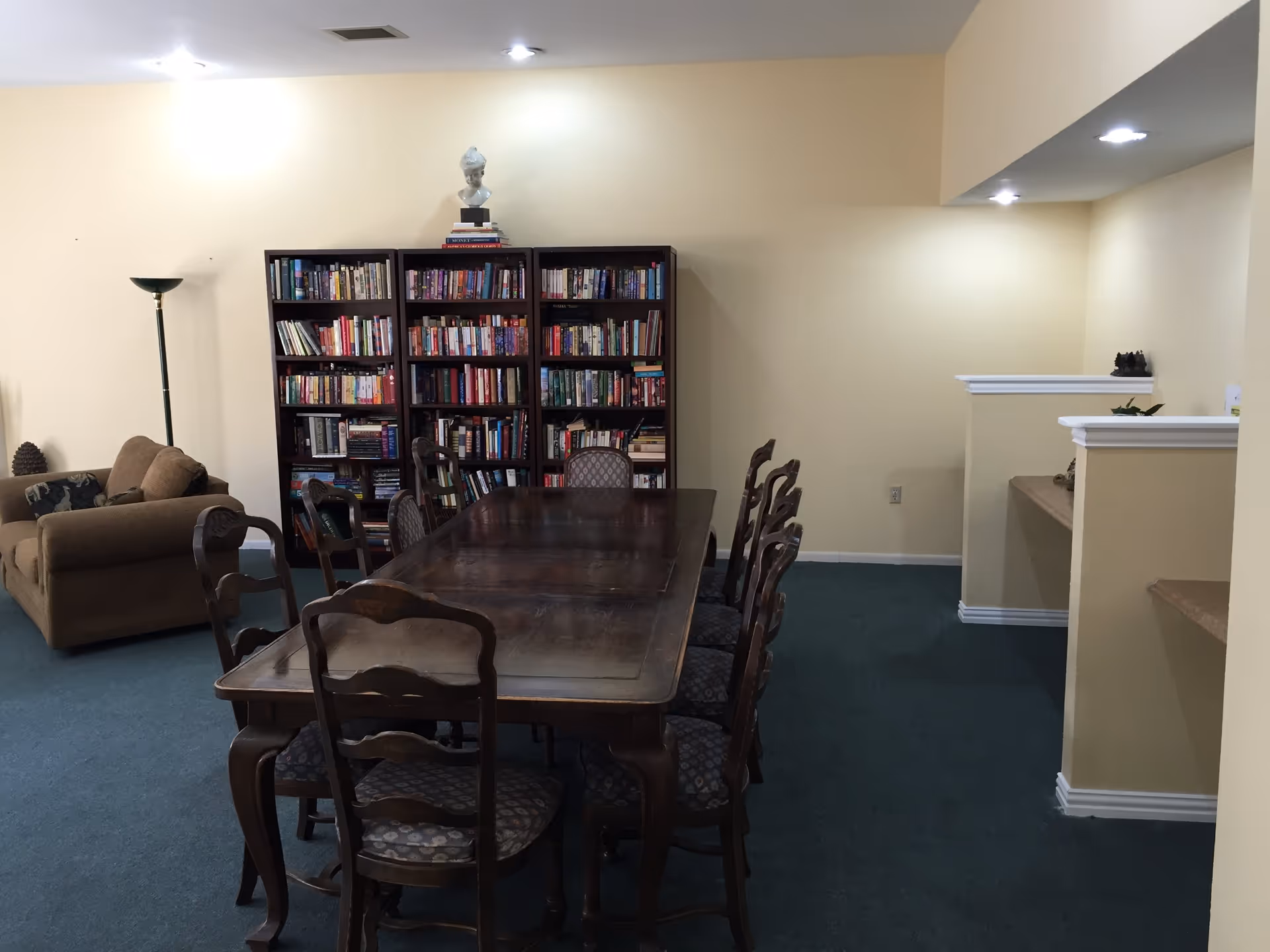 A room with a long wooden table surrounded by eight chairs with patterned cushions. Behind the table is a large bookshelf filled with books and topped with a bust sculpture. To the left, there is a brown sofa and a tall floor lamp. The walls are painted light beige and the floor is covered with dark green carpet. The room is lit by ceiling lights.