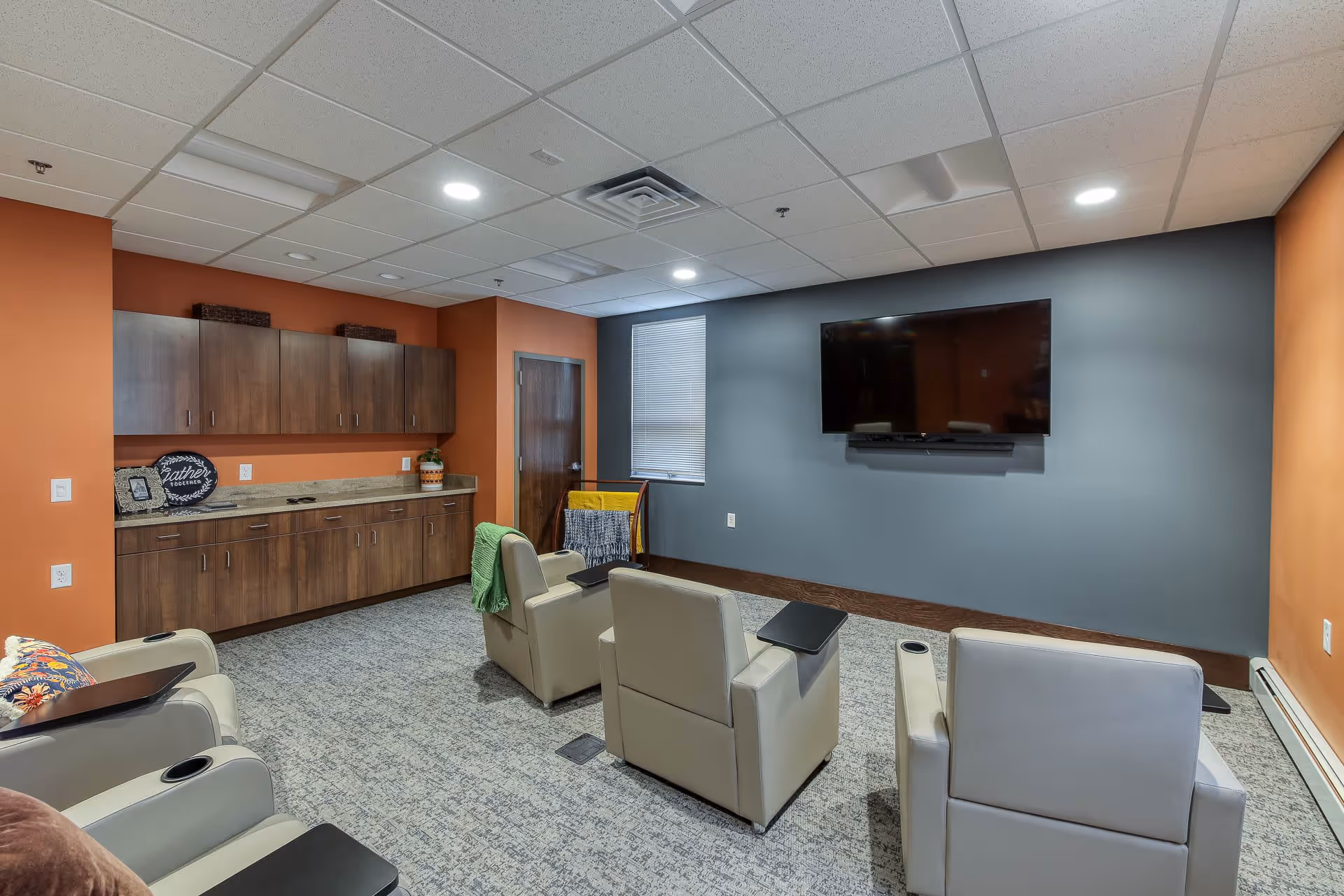 A cozy senior living common room with four beige recliner chairs facing a large flat-screen TV mounted on a gray wall. The room has orange accent walls, a carpeted floor, and wooden cabinets with a countertop along one side. A small window with blinds is next to a wooden door, and a blanket rack with colorful blankets is placed near the window.