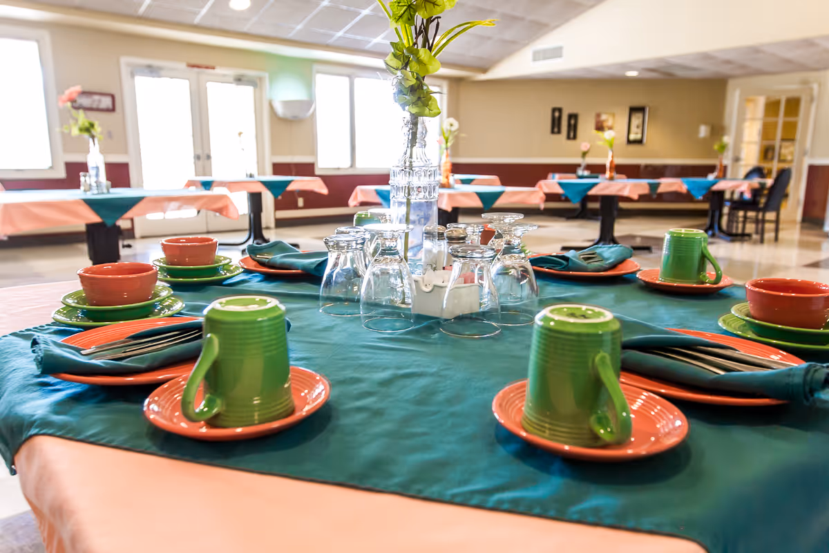 A dining room set up with tables covered in peach and teal tablecloths, each table set with green mugs, orange plates, bowls, and silverware. The room has large windows and double doors letting in natural light, with additional tables and chairs visible in the background.