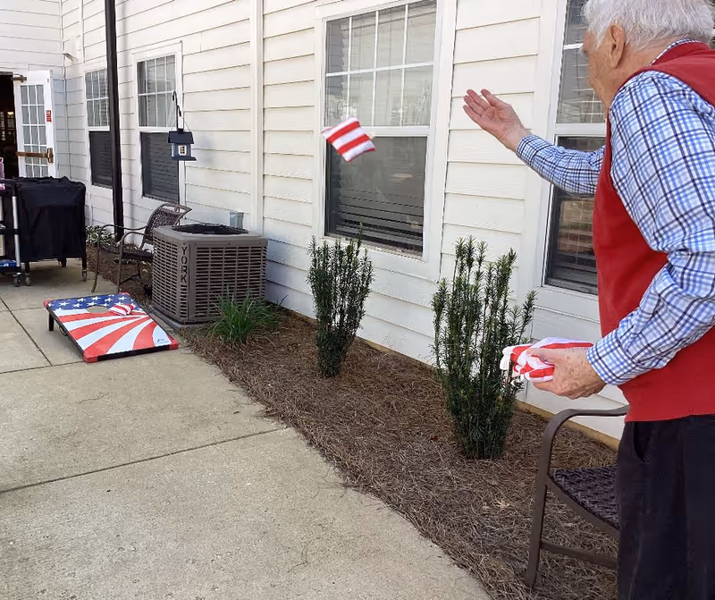 An elderly man wearing a red vest and blue checkered shirt is playing a bean bag toss game outside next to a white building with windows. The bean bag board has an American flag design, and the man is throwing a red and white striped bean bag towards it. There are small bushes planted along the building and a chair nearby.