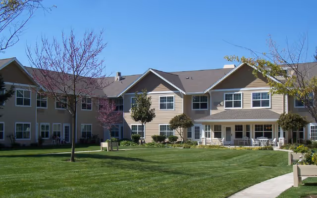 Exterior view of a two-story senior living facility building with beige siding and white trim, surrounded by a well-maintained green lawn, small trees, and outdoor seating areas under a clear blue sky.