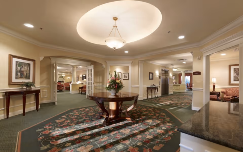 Well-lit senior living facility lobby with a central round table topped by a floral arrangement and seating areas beyond.