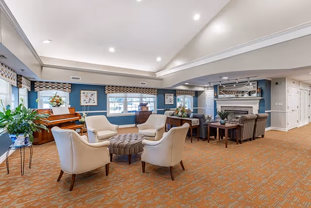 A spacious and well-lit living room area with beige armchairs arranged around a round ottoman, a piano near the windows, a fireplace with a mantel, and various plants and decorative items. The walls are painted blue with white trim, and the floor is covered with a patterned carpet.